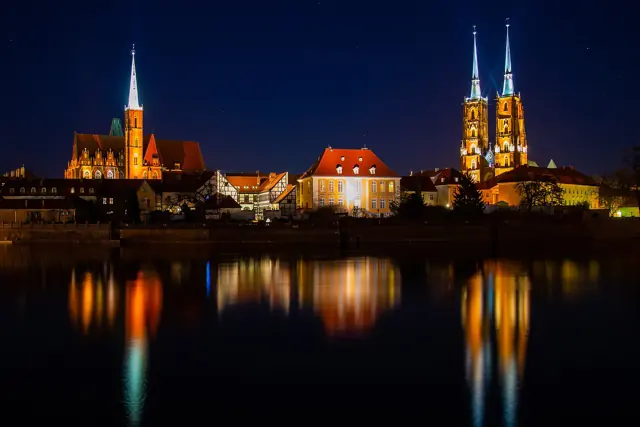 The towers of Wrocław Cathedral and the Holy Cross church on Ostrów Tumski, lit at night and reflected in the Oder